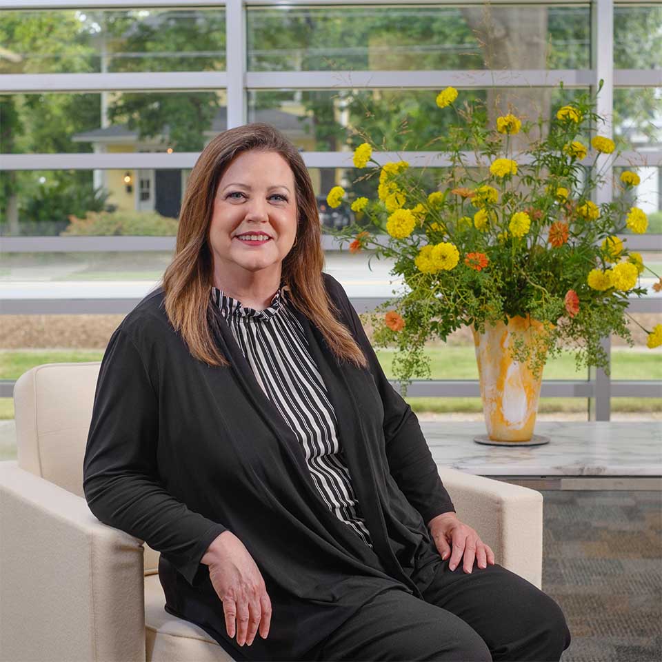 Susan McCants seated in front of floor-to-ceiling windows and a vase with yellow and orange flowers.