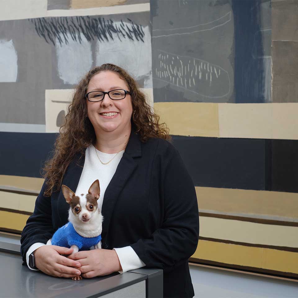 Jessica Daley standing at a counter with a chihuahua in a sweater in front of an abstract wall mural.