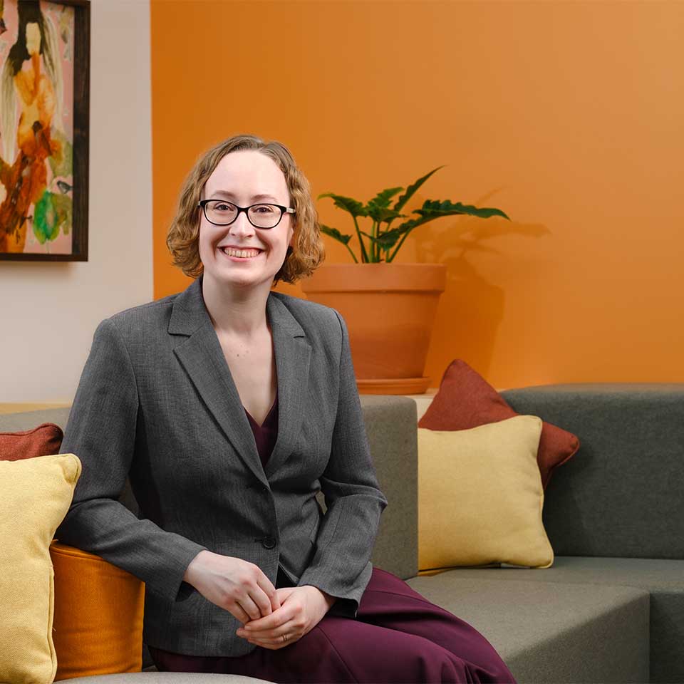 Anne Serrano seated on a grey couch in front of an orange wall with abstract art and a potted plant in the background.